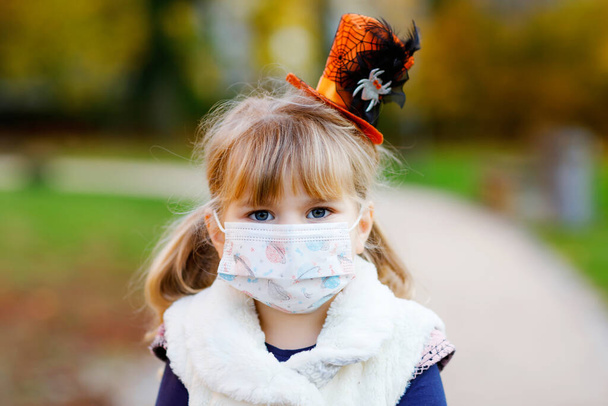 Κοριτσάκι νήπιο ντυμένο μάγισσα με ιατρική μάσκα στο πρόσωπο. Παιδί σε εξωτερικούς χώρους, κατά τη διάρκεια της καραντίνας του ιού Πανδημίας με προστατευτικό εξοπλισμό - Φωτογραφία, εικόνα