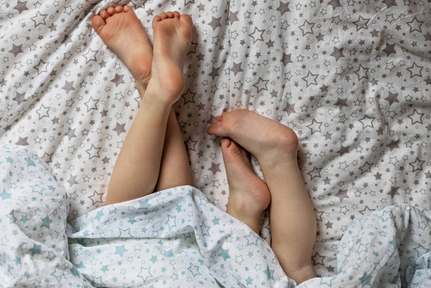 Nahaufnahme von zwei Paar kleinen Mädchen Füße auf dem Bett unter der Decke. Helle Blau- und Beigetöne - Foto, Bild