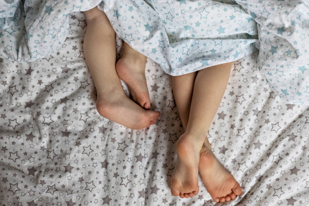 Close-up van twee paar peutermeisjes voeten op het bed onder de deken. Lichtblauwe en beige tinten - Foto, afbeelding
