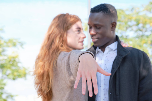 アフリカ系黒人男性と赤毛の白人女性がカメラに婚約指輪を見せ、お互いを見ていた。婚姻の建議. - 写真・画像