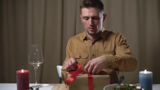 un homme s'assoit à une table festive et ouvre une boîte avec un cadeau. Détache le ruban de satin rouge. Le concept de cadeaux pour les proches le jour de l'anniversaire, la Saint-Valentin. - Séquence, vidéo