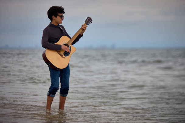 young caucasian teenager playing acoustic guitar on the beach in the water. sunglasses and dark clothing - Фото, изображение