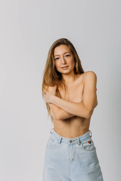 a young beautiful girl with dyne loose hair topless in jeans in the studio on a white background makes different emotions on her face holding and covering her breasts with her hands - Photo, Image