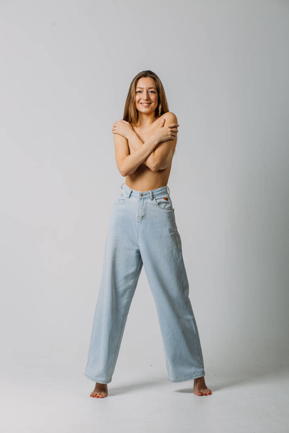 a young beautiful girl with dyne loose hair topless in jeans in the studio on a white background makes different emotions on her face holding and covering her breasts with her hands - Photo, Image