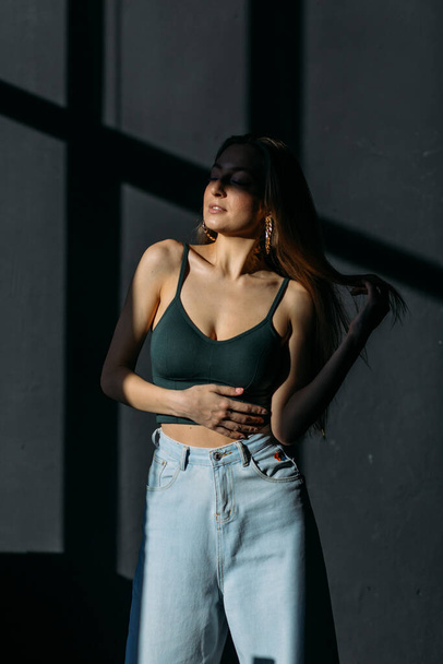young beautiful girl with dyne loose hair in a top and jeans in the studio on a black background in the rays of the sun - Photo, Image