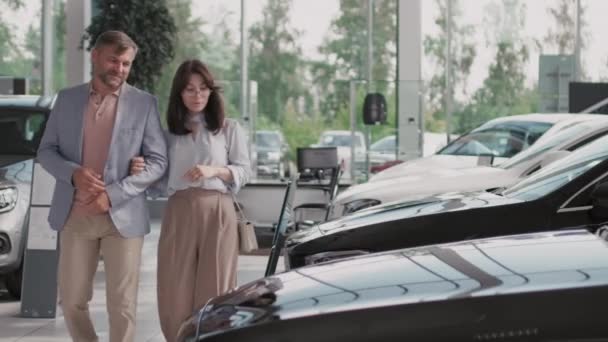 Keskikokoinen laukaus älykkäästi pukeutunut pari kävelee yhdessä autokaupassa ja puhuu valitessaan ajoneuvoa - Materiaali, video