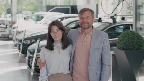 Tracking portrait shot of smartly dressed young woman and middle-aged man standing together in car dealership and posing for camera - Footage, Video