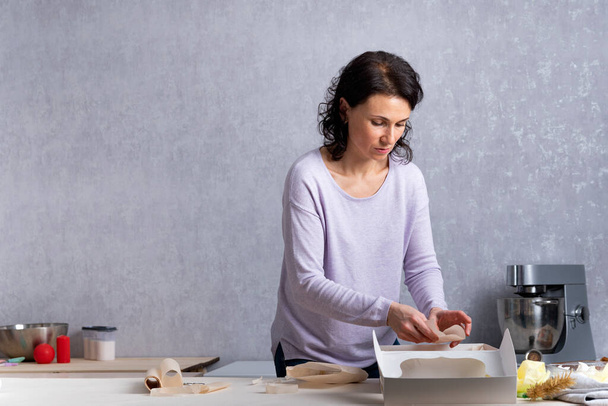 Young woman in kitchen is packing pastries in white cardboard box. Process of wrapping . - Photo, Image