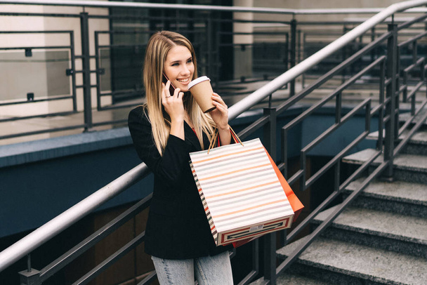 Portrait d'une belle jeune fille qui sourit en parlant au téléphone après avoir fait du shopping avec du café dans les mains et des sacs à provisions. - Photo, image