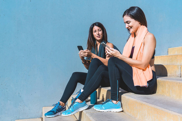 twee jonge vrouwen dragen sportkleding en zitten op een trap en nemen een pauze en controleren hun smartphones. Begrip sport en technologie - Foto, afbeelding