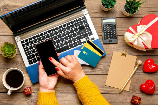 Girl holds smartphone, chooses gift, makes purchases, debit card, laptop, cup of coffee, two hearts on wooden table Top view Flat lay Holiday shopping list, Happy Valentines day, online shop concept - Foto, Imagen