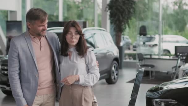 Slowmo PAN toma de feliz joven mujer y su marido de mediana edad de pie juntos en la concesionaria y hablando al elegir un coche nuevo - Imágenes, Vídeo