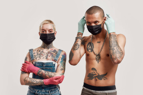 Quarantine accessories. A white woman and a dark-skinned man both tattooed wearing black respiratory masks and rubber gloves - 写真・画像