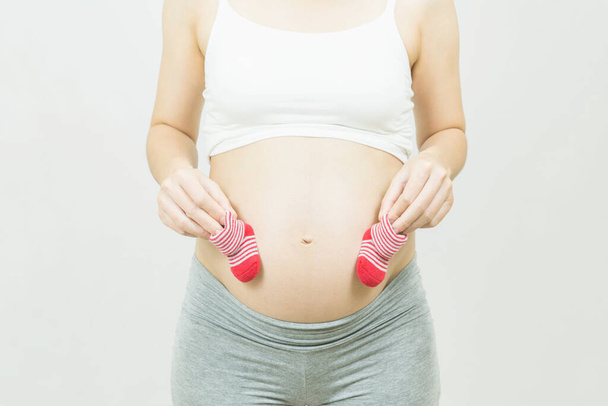 A tummy of a pregnant woman with red baby's bootees,Pregnancy health care preparing for baby concept.Motherhood among teenage mother, hugging belly, isolated on gray background - Valokuva, kuva