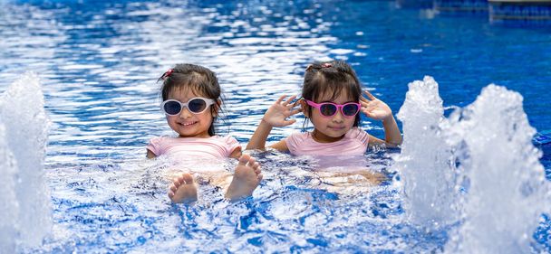 Ritratto di graziosi gemelli asiatici che sorridono e posano sullo sfondo della piscina indossando costume da bagno rosa e occhiali da sole - Foto, immagini