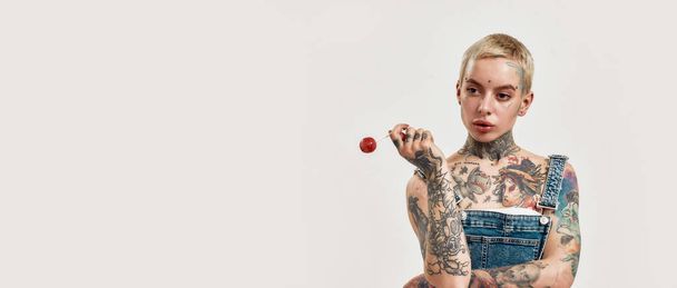 Tatuaje y piercing. Una mujer blanca perforada con tatuajes con un denim en general sosteniendo una piruleta roja mientras mira hacia los lados - Foto, imagen