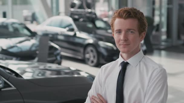 Retrato lento del joven vendedor de coches con corbata y camisa blanca de pie con los brazos cruzados y mirando a la cámara en el concesionario - Imágenes, Vídeo