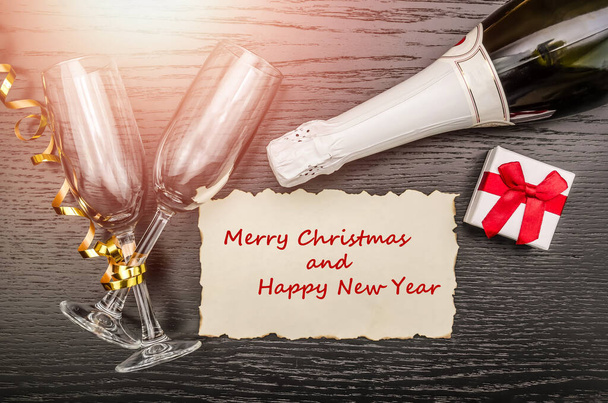 Two glasses, a bottle of champagne, a gift box, a sheet with the inscription Merry Christmas and Happy New Year on a dark background with lighting, top view from copy space. Concept Of Christmas - Photo, Image