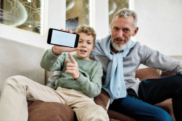 Cute little boy pointing on blank mobile phone screen, while relaxing on sofa with his grandfather at home - Foto, Imagem