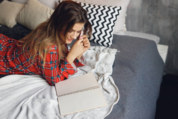 Top view of girl wears festive pajamas lies and write Wish List at home. Smiling woman in red night-wear on bed - Photo, image