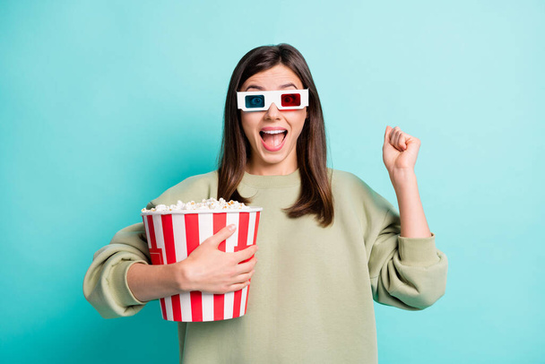 Photo portrait of screaming girl in 3d glasses holding large popcorn bucket with raised fist isolated on vivid turquoise colored background - Foto, afbeelding