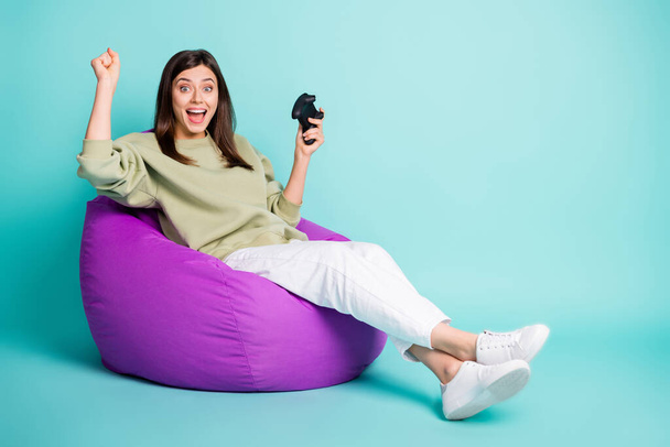 Photo portrait full body view of excited woman celebrating victory in game holding controller in one hand sitting in purple chair isolated on vivid cyan colored background - Fotó, kép