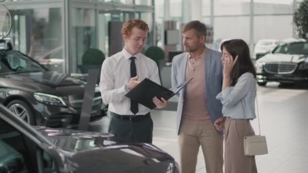Tracking of young dealership salesman in formal attire holding folder and showing car documentation to couple shopping for new vehicle - Footage, Video