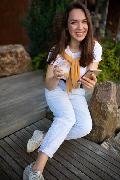 young woman freelancer sits in the park and enjoys a coffee break with a phone in her hands - Foto, Imagem