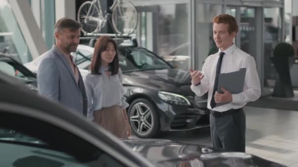 Tracking shot of young car salesman with clipboard talking to couple in dealership, then shaking hand of man - Footage, Video