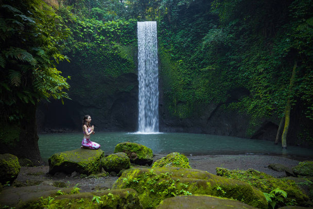 Nuori valkoihoinen nainen istuu kivellä lähellä vesiputousta Vajrasanassa tai Diamond poseeraa. Kädet namumaisessa mudassa. Trooppinen näkymä. Jooga ja pranayama käsite. Rentouttava tunnelma. Tibumanan vesiputous, Bali - Valokuva, kuva