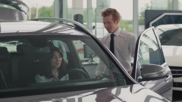 Tracking shot of cheerful young salesman in formal attire walking up to beautiful young woman sitting inside expensive car in dealership and answering her questions - Footage, Video