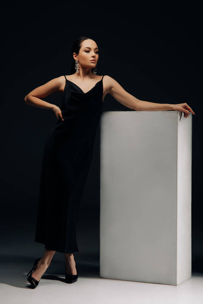 Junge Frau posiert in langem schwarzen Kleid und Stöckelschuhen am Rednerpult vor schwarzem Hintergrund. - Foto, Bild