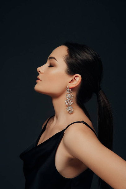 Portrait of young woman in black dress with earrings and makeup on her face against of black background. Side view. - Photo, Image