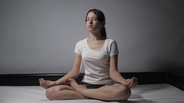 Zrelaksowana młoda kobieta w piżamie medytuje w pozycji lotosu siedząc na łóżku z zamkniętymi oczami w domu i medytując rękami. Dziewczyna chodzi rano na gimnastykę. Koncepcja zdrowia i spokoju - Materiał filmowy, wideo