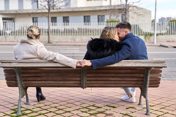 Love triangle, a girl kiss at her boyfriend and he shakes hands with another girl, they are sitting on a bench. Concept of Infidelity - Photo, Image