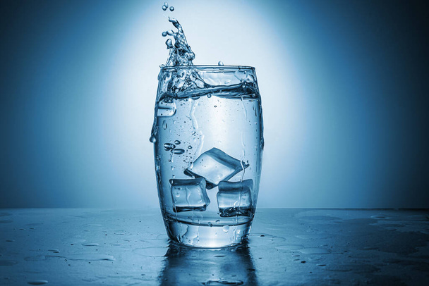 Un vaso de agua con cubitos de hielo. Salpicadura de H2o en un recipiente transparente. Sombra azul. - Foto, imagen
