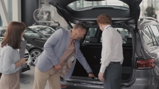 Tracking shot of young salesman in formal attire showing boot of car to cheerful couple in dealership. He is answering their questions and smiling - Footage, Video
