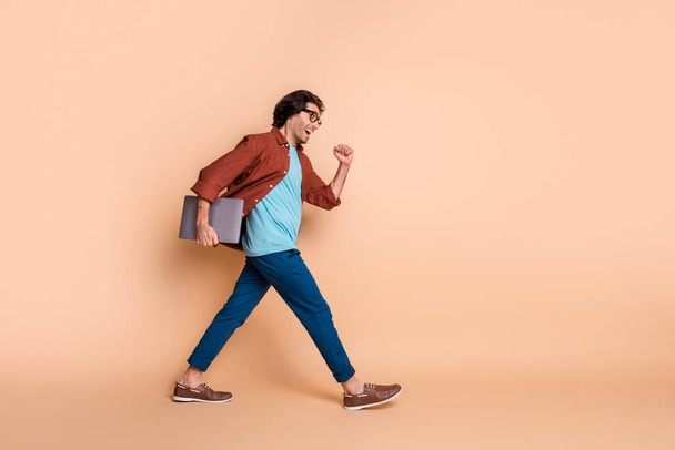 Full length profile photo of positive man hold laptop walk fast wear eyeglasses brown t-shirt isolated beige color background - Fotoğraf, Görsel