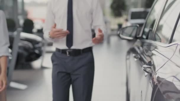 Παρακολούθηση με τη μέση του τμήματος αγνώριστη νεαρός πωλητής στην επίσημη ενδυμασία με τα πόδια και μιλώντας με τη γυναίκα πελάτη, στη συνέχεια, ανοίγοντας την πόρτα του αυτοκινήτου για να της δείξει το εσωτερικό του - Πλάνα, βίντεο