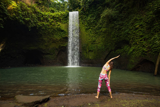 Biała kobieta ćwiczy jogę przy wodospadzie. Trikonasana, Trójkąt Pose. Stojąca Asana. Wspieraj układ odpornościowy. Odwrót jogi. Koncepcja zdrowia. Widok z tyłu. Wodospad Tibumana, Bali - Zdjęcie, obraz