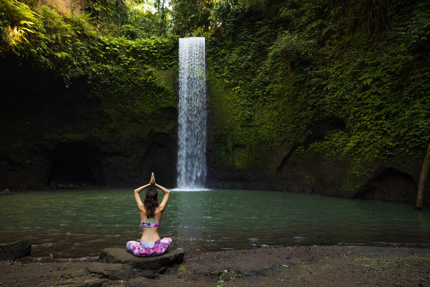 Поза йоги лотоса. Молодая кавказка сидит на камне, медитирует на йоге, пранаяма у водопада. Руки подняты в мудре намасте. Отдых на йоге. Вид сзади. Тибуманский водопад - Фото, изображение