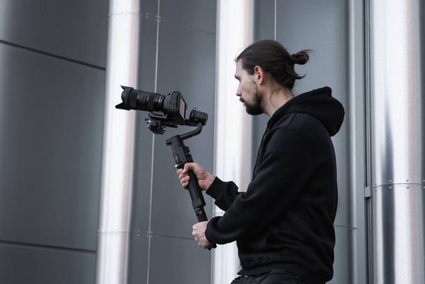 Young Professional videographer holding professional camera on 3-axis gimbal stabilizer. Pro equipment helps to make high quality video without shaking. Cameraman wearing white hoodie making a videos. - Foto, afbeelding