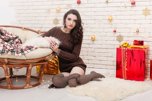 Beautiful curly brunette girl at the Christmas tree with toys, decorations, lights. Young woman in a warm knitted sweater, long golf leggings holding a gift box in her hands, smiling by fireplace - Φωτογραφία, εικόνα