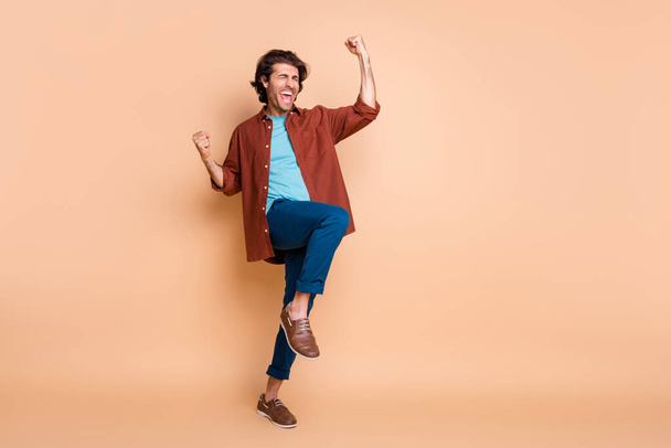 Full length photo of crazy man raise fists open mouth wear brown t-shirt blue trousers shoes isolated beige color background - 写真・画像