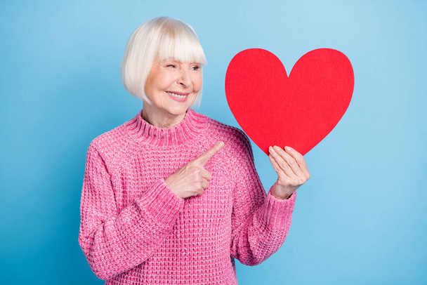 Foto retrato de mulher velha olhando apontando dedo para o grande cartão de coração vermelho segurando em uma mão isolada no fundo de cor azul pastel - Foto, Imagem