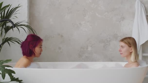 Young beautiful Asian and Caucasian women sitting together in bathtub, looking at each other and then posing for camera - Footage, Video