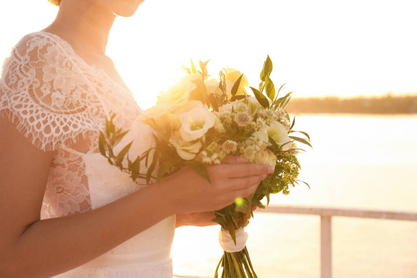 Bride in beautiful wedding dress with bouquet near river on sunset, closeup - Foto, Imagem