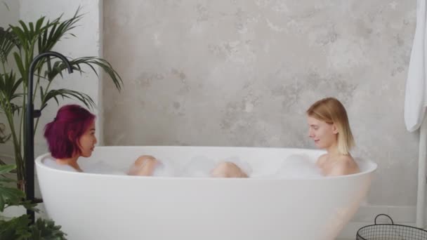 Jonge mooie Aziatische en blanke vrouwelijke vrienden zitten samen in bad, spelen met schuim en chatten - Video