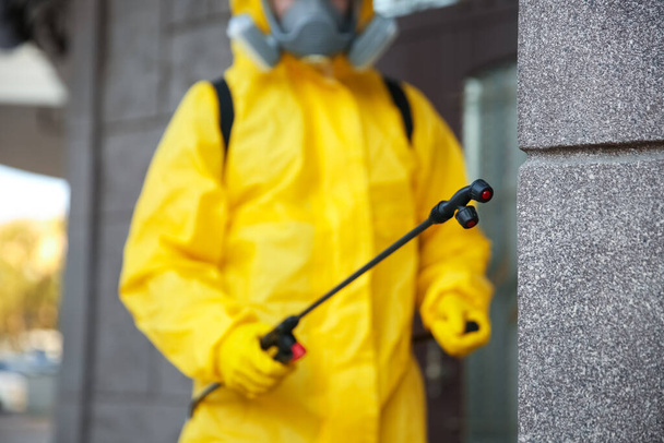 Person in hazmat suit disinfecting building wall outdoors, focus on sprayer. Surface treatment during coronavirus pandemic - Фото, зображення