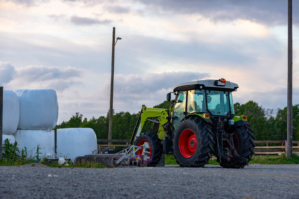Фермерский трактор возле собранных тюков сена, завернутых в пластик на фермерском поле на закате возле леса, солнечный теплый свет. - Фото, изображение